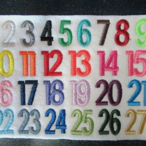 Puede incluir: Un cuadrado de fieltro blanco bordado con n&uacute;meros coloridos del 1 al 27. Los n&uacute;meros son de varios colores, incluyendo plata, negro, verde, rojo y azul. Los n&uacute;meros est&aacute;n en una fuente simple y clara.