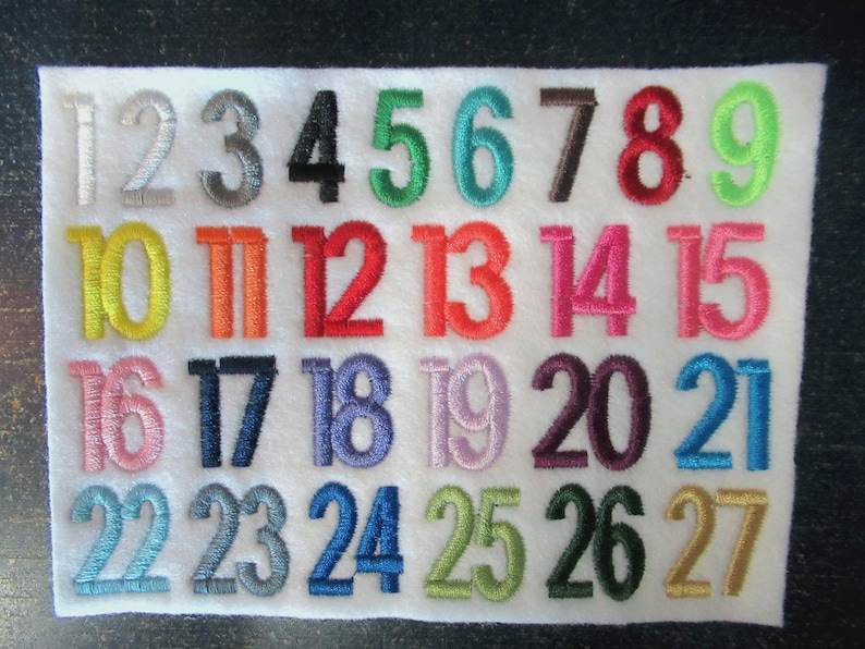 Puede incluir: Un cuadrado de fieltro blanco bordado con n&uacute;meros coloridos del 1 al 27. Los n&uacute;meros son de varios colores, incluyendo plata, negro, verde, rojo y azul. Los n&uacute;meros est&aacute;n en una fuente simple y clara.