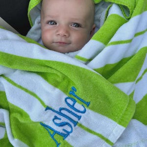 May include: A baby wrapped in a green and white striped towel with the name "Asher" embroidered in blue. The towel has a hood. The baby is looking at the camera.