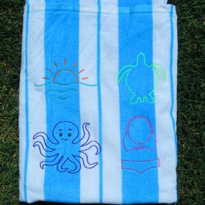 Puede incluir: Toalla de playa a rayas azules y blancas con dise&ntilde;os bordados. Presenta un sol, una tortuga, un pulpo y un bikini. La toalla est&aacute; doblada y sobre hierba verde.