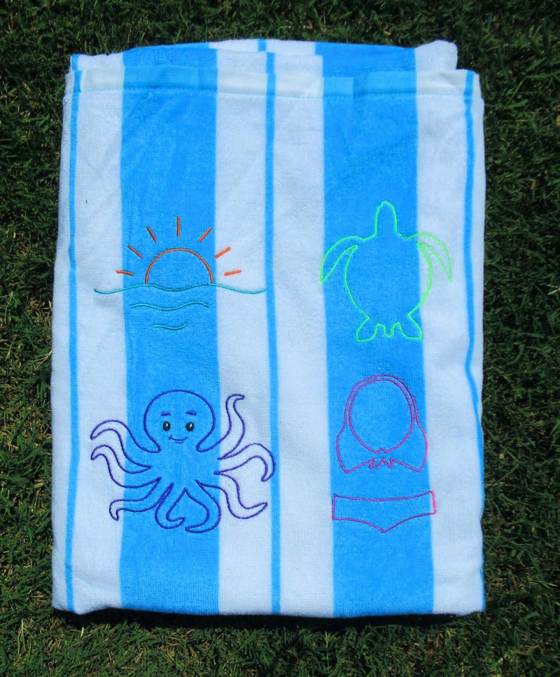 Puede incluir: Toalla de playa a rayas azules y blancas con dise&ntilde;os bordados. Presenta un sol, una tortuga, un pulpo y un bikini. La toalla est&aacute; doblada y sobre hierba verde.