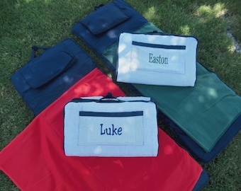 May include: Picnic blankets in red, green, and navy blue are displayed on grass. Two blankets have a blue and white striped pattern with the names "Easton" and "Luke" embroidered on them. The blankets are folded and have handles.