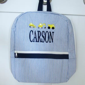 May include: Blue and white striped backpack with a white zipper and navy blue accents. The backpack features embroidered yellow construction vehicles and the name "CARSON" in navy blue.