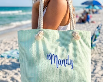 May include: A light green and white seersucker tote bag featuring thick white rope handles with knotted ends and the name Mandy embroidered in dark blue script lettering on the front centre of the bag, providing a textured fabric appearance.
