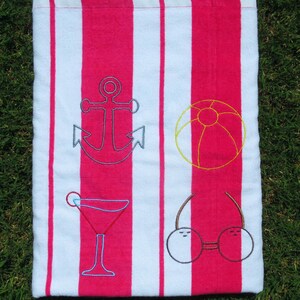 Puede incluir: Una toalla de playa con rayas verticales alternas blancas y rosa brillante. Bordados en la toalla hay un ancla azul, un bal&oacute;n de playa amarillo, un c&oacute;ctel rojo y unas gafas de sol con cerezas.