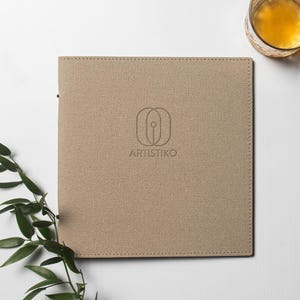 Pode incluir: Um caderno quadrado, cor de bege, com a palavra "ARTISTIKO" impressa na capa, ao lado de um logotipo. Um copo de líquido âmbar está à direita e um ramo de folhas verdes à esquerda.