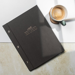 Può includere: Un menu o taccuino grigio scuro con un logo dorato in rilievo e rivetti argentati. Una tazza di caffè è appoggiata su un sottobicchiere di legno. Il testo sulla copertina recita "THE WILDERNESS HOTEL".