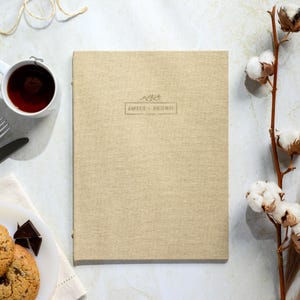 Könnte beinhalten: Ein beiges Buch mit Leinenstruktur und dem Text "EMILY + BRUNO" in einem rechteckigen Rahmen. Das Bild zeigt eine weiße Teetasse mit Tee, einen Teller mit Keksen, Besteck, Baumwollstiele und Bindfaden.