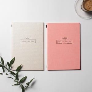 Peut inclure: Deux carnets à couverture rigide, un beige et un rose, sont présentés sur une surface blanche. Chaque carnet porte l'inscription "EMILY + BRUNO" dans un cadre rectangulaire avec un motif floral. Une tasse de café est à proximité.
