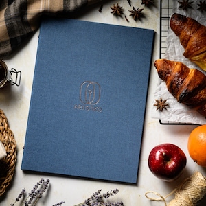 Może przedstawiać: Niebieskie menu w oprawie lnianej z napisem "Artistico" złotymi literami. Menu leży na białym tle z różnymi produktami spożywczymi, w tym croissant, jabłkiem, pomarańczą i anyżem gwiazdkowym.