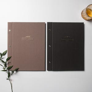 Może przedstawiać: Dwie oprawione książki, jedna brązowa i jedna czarna, ze srebrnymi okuciami. Każda książka ma wytłoczone złotym logo i tekst. W pobliżu stoi szklanka z bursztynowym płynem. Gałązka zielonych liści znajduje się w lewym dolnym rogu.