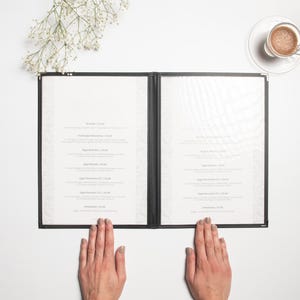 Könnte beinhalten: Ein offenes schwarzes Menü mit weißen Seiten, gehalten von zwei Händen. Rechts steht eine Tasse Kaffee auf einer Untertasse, und links oben befindet sich ein Zweig mit weißen Blüten. Das Menü enthält Text, wahrscheinlich eine Liste von Speisen.