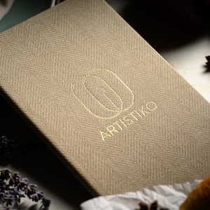 Może przedstawiać: Beige'owa okładka menu z lnu z wytłoczonym złotym logo z napisem "Artistico" i geometrycznym wzorem.