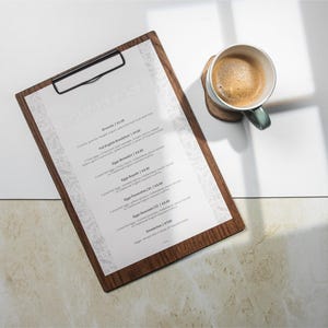 Könnte beinhalten: Ein hölzernes Klemmbrett mit einer gedruckten Frühstückskarte, die eine Liste von Gerichten und Preisen enthält. Eine Tasse Kaffee mit hellbrauner Crema steht auf einem Untersetzer aus Holz. Die Szene wird von natürlichem Licht beleuchtet.