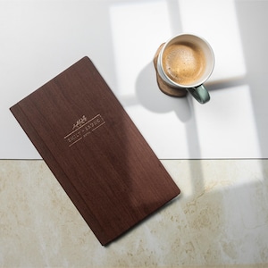 Könnte beinhalten: Ein dunkelbraunes Holzmenü mit dem Text "Emily & Ringo" und eine Kaffeetasse mit Untertasse. Der Kaffee hat eine hellbraune Crema. Die Tasse ist türkis und weiß.