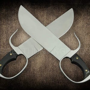 May include: Two crossed, silver-bladed butterfly swords with black handles and finger loops. The blades are wide and curved, and the handles are riveted with gold-colored studs. The swords are set against a brown background.