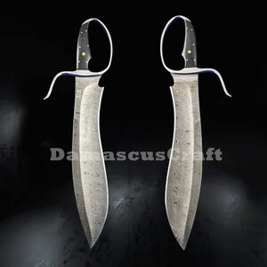 Op de afbeelding: Twee Damascus stalen messen met gebogen lemmeten en open lus handvatten. De handvatten zijn zwart met gouden accenten. De lemmeten hebben een uniek patroon. De tekst "Damascuscraft" is zichtbaar.