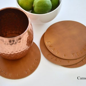 May include: Set of four brown leather coasters with the names "Evan & Tessa" embossed on each. The coasters are arranged in a circle with a copper mug on top of one of the coasters. The background is white.