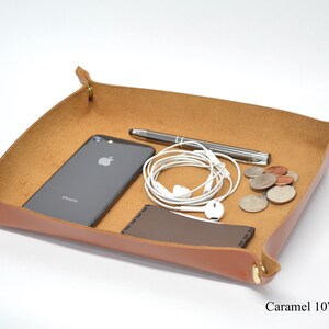 Puede incluir: Una bandeja de cuero caramelo con detalles en lat&oacute;n, que incluye un tel&eacute;fono inteligente, auriculares, un bol&iacute;grafo y monedas. La bandeja mide 25,4 cm por 20,3 cm.