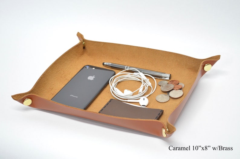 Puede incluir: Una bandeja de cuero caramelo con detalles en lat&oacute;n, que incluye un tel&eacute;fono inteligente, auriculares, un bol&iacute;grafo y monedas. La bandeja mide 25,4 cm por 20,3 cm.