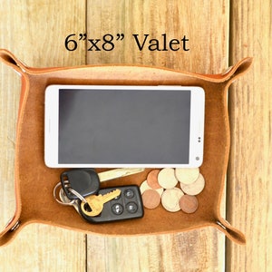 Puede incluir: Una bandeja valet de cuero marr&oacute;n con un tel&eacute;fono inteligente, llaves y monedas en su interior. La bandeja mide 15,24 cm por 20,32 cm.