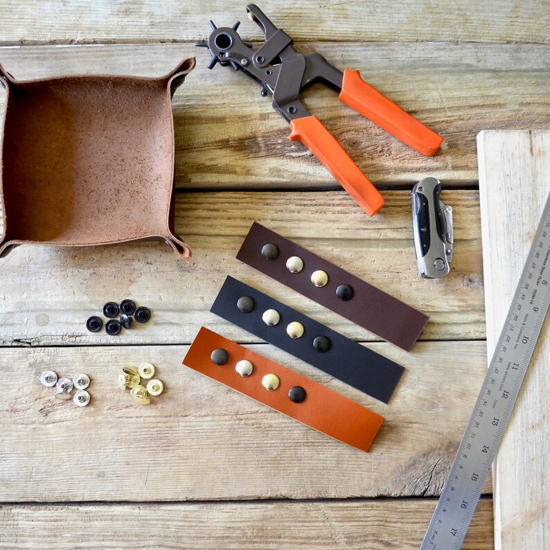 Puede incluir: Primer plano de un proyecto de artesan&iacute;a de cuero con tres tiras de cuero, cada una con remaches de diferentes colores. Las tiras de cuero son marr&oacute;n, negro y naranja. Tambi&eacute;n hay herramientas para la artesan&iacute;a del cuero, incluyendo un punz&oacute;n, una regla y un cuchillo multiusos.