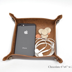 Puede incluir: Una bandeja de cuero marr&oacute;n con remaches de lat&oacute;n antiguo. La bandeja mide 15,24 cm por 15,24 cm y est&aacute; llena de un tel&eacute;fono, auriculares y monedas.