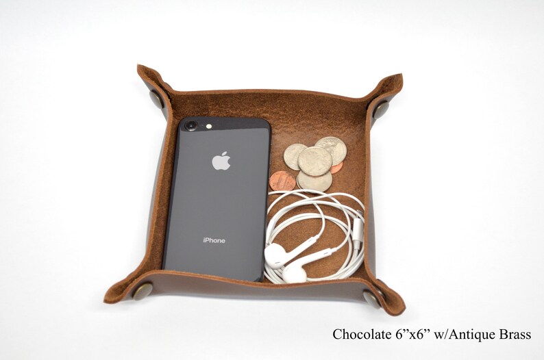 Puede incluir: Una bandeja de cuero marr&oacute;n con remaches de lat&oacute;n antiguo. La bandeja mide 15,24 cm por 15,24 cm y est&aacute; llena de un tel&eacute;fono, auriculares y monedas.