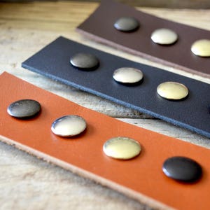 Puede incluir: Tres correas de cuero con remaches met&aacute;licos de diferentes colores. Las correas son marr&oacute;n, negra y naranja. Los remaches son plateados, dorados y negros.