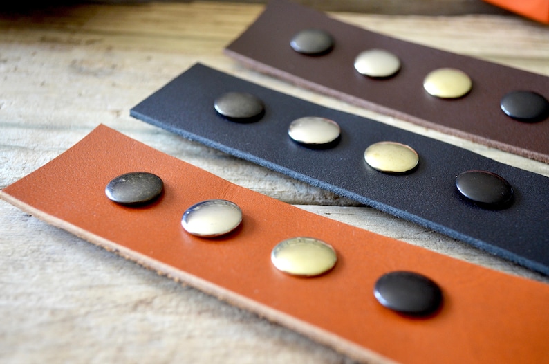 Puede incluir: Tres correas de cuero con remaches met&aacute;licos de diferentes colores. Las correas son marr&oacute;n, negra y naranja. Los remaches son plateados, dorados y negros.