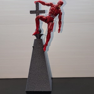 May include: A red action figure of Daredevil standing on a black pyramid-shaped base with a black cross on top. The figure is in a dynamic pose, with one leg raised and one arm extended.