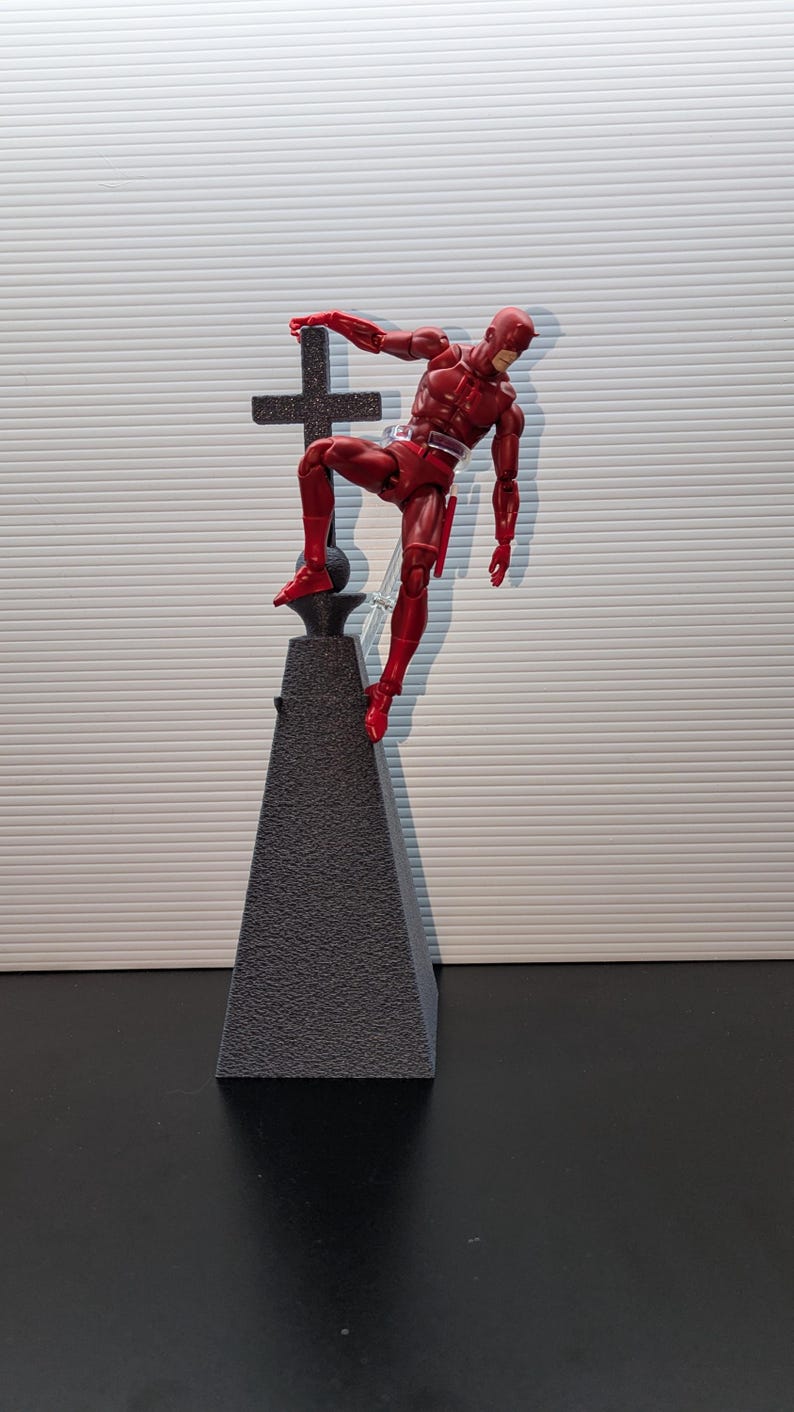 May include: A red action figure of Daredevil standing on a black pyramid-shaped base with a black cross on top. The figure is in a dynamic pose, with one leg raised and one arm extended.