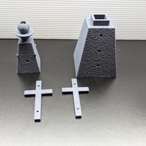 May include: A set of gray 3D printed miniature cemetery decorations. The set includes two small crosses, a small obelisk with a sphere on top, and a larger obelisk with a square top.