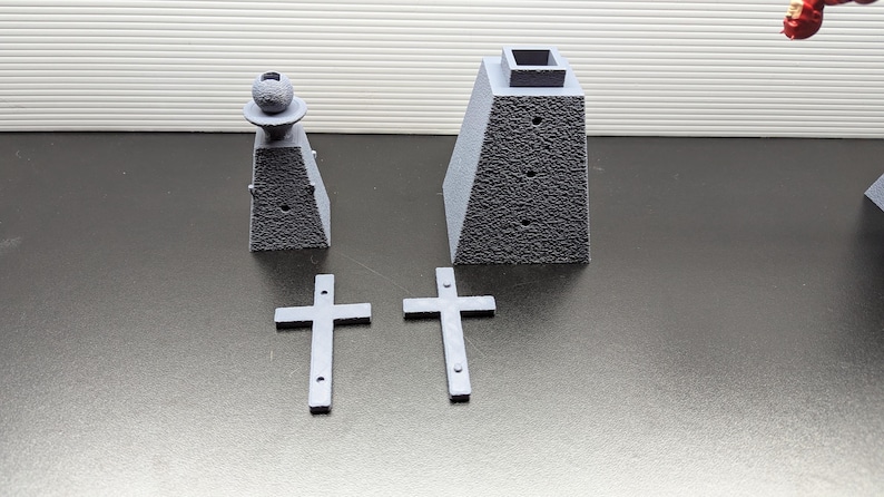 May include: A set of gray 3D printed miniature cemetery decorations. The set includes two small crosses, a small obelisk with a sphere on top, and a larger obelisk with a square top.