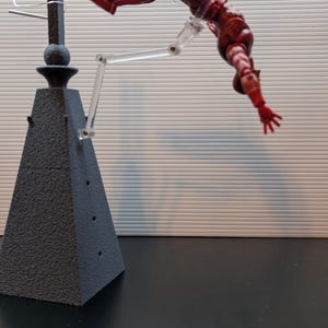 May include: A red action figure of a superhero is suspended in mid-air by a clear plastic stand. The figure is posed in a dynamic, action-packed pose, with one arm extended and the other bent at the elbow. The figure is attached to a black, pyramid-shaped base with a cross on top.