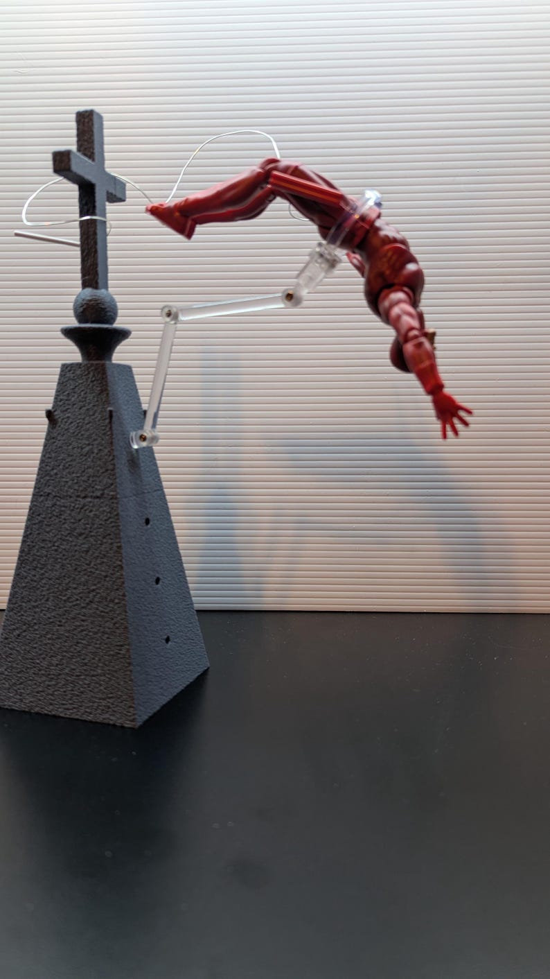 May include: A red action figure of a superhero is suspended in mid-air by a clear plastic stand. The figure is posed in a dynamic, action-packed pose, with one arm extended and the other bent at the elbow. The figure is attached to a black, pyramid-shaped base with a cross on top.