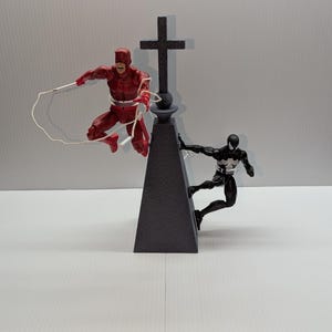 May include: A red and black action figure of Daredevil is swinging a white rope towards a black action figure of Spider-Man, who is climbing a gray pyramid-shaped structure with a black cross on top.