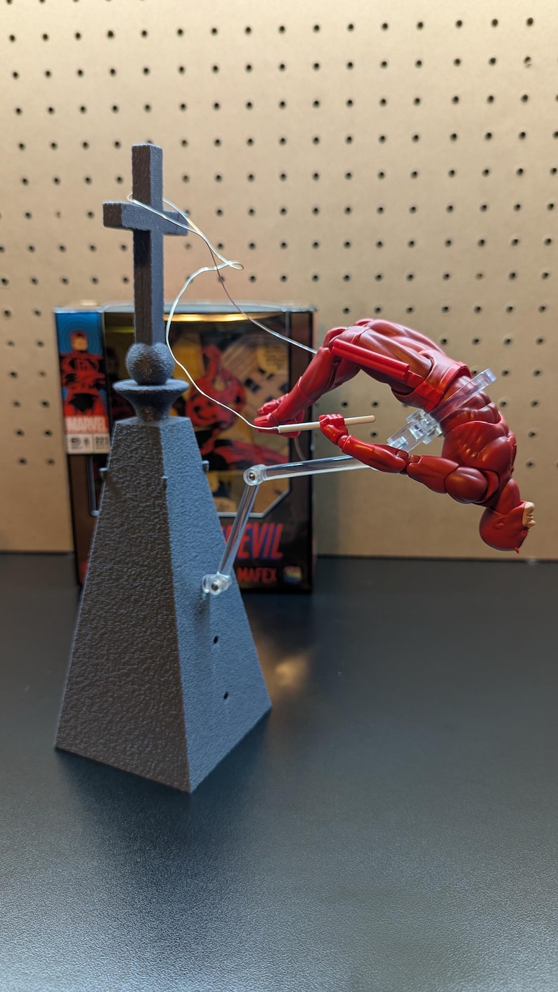 May include: A red Daredevil action figure is suspended in mid-air by a clear plastic stand. The figure is posed in a dynamic action pose, with one leg extended and the other bent. The figure is holding a stick in its right hand. The figure is attached to a gray base with a cross on top. The base has the words "Marvel" and "MAFEX" printed on it.