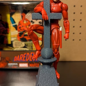 May include: A red Daredevil action figure standing on a gray stone base with a cross. The figure is holding onto the cross with one hand and has a red stick in the other hand. The figure is wearing a red suit with a red mask. The base is textured and has a round top.