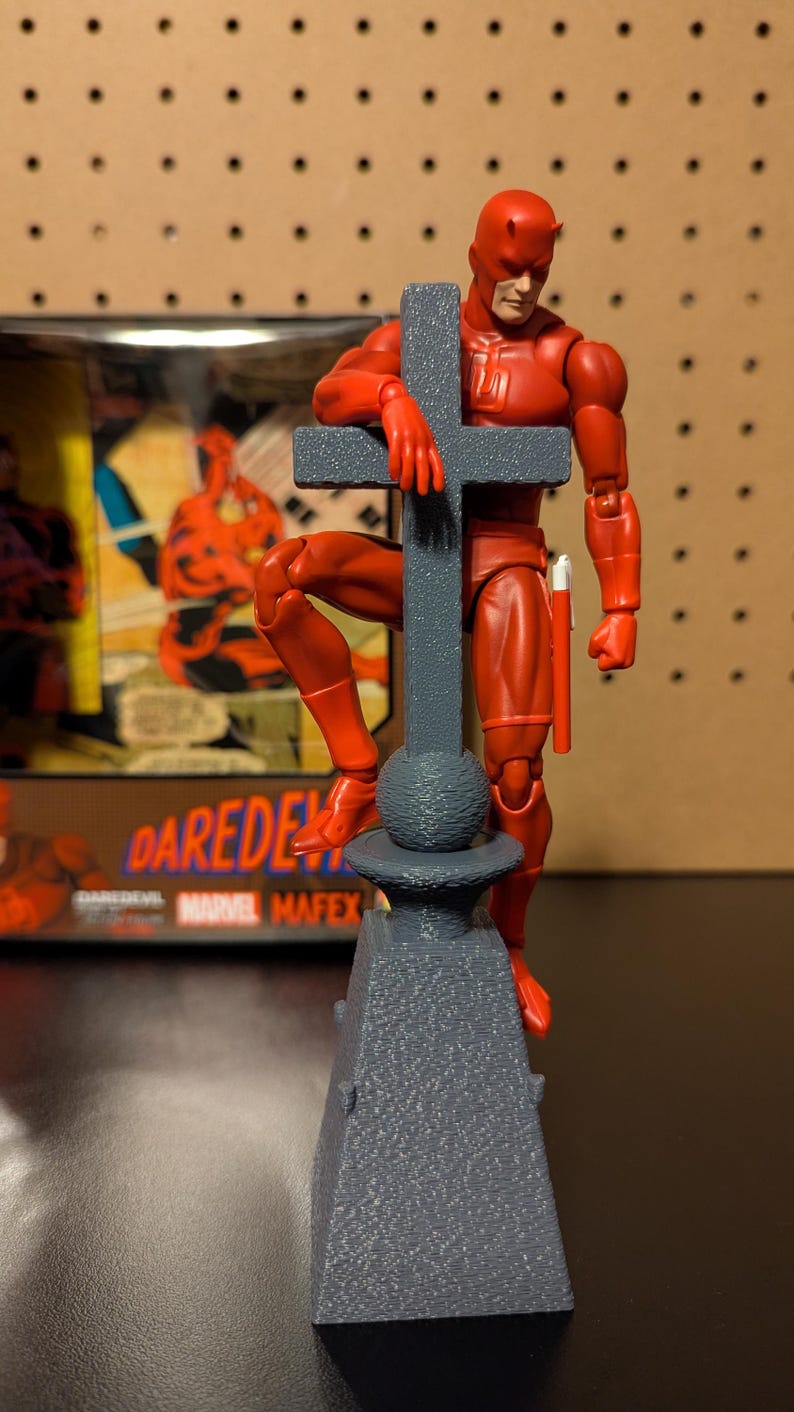 May include: A red Daredevil action figure standing on a gray stone base with a cross. The figure is holding onto the cross with one hand and has a red stick in the other hand. The figure is wearing a red suit with a red mask. The base is textured and has a round top.