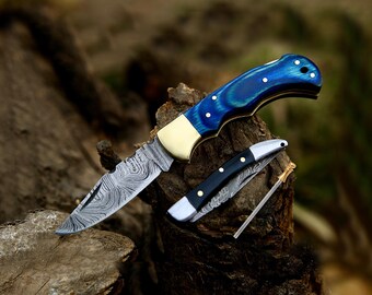 May include: A large folding knife with a blue and gold handle and a smaller black-handled knife are displayed on a piece of wood. The blades have a unique patterned design. A small, gold-tipped tool is also present.
