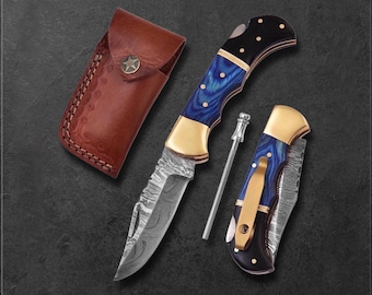 May include: A set of three knives with a brown leather sheath. The knives have blue and black handles with gold accents. The blades have a unique pattern. The sheath has a star-shaped button.