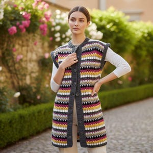 May include: A sleeveless, knee-length crochet vest featuring a button-down front with a dark gray ribbed placket. The garment displays a repeating horizontal striped pattern in shades of magenta, mustard yellow, white, and black throughout.
