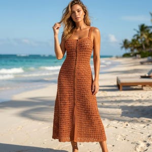 May include: A rust-colored, sleeveless midi-length dress crafted from a crocheted knit fabric. The garment features a straight neckline with thick shoulder straps and a vertical row of small buttons running down the center front from the bodice to the hem.