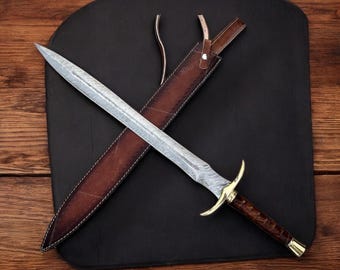 May include: A double-edged dagger with a patterned Damascus steel blade, a polished brass crossguard, and a dark brown textured handle. The dagger is accompanied by a matching brown leather sheath featuring white stitching and a strap closure.