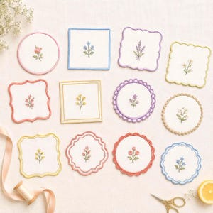 May include: A collection of twelve white fabric coasters with embroidered floral motifs in various colours. The coasters feature diverse shapes including circles, squares, and scalloped edges with matching coloured borders in pink, blue, yellow, and purple.