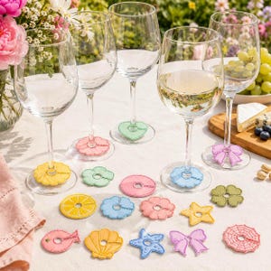 May include: A collection of clear glass wine glasses and assorted felt wine glass charms in various shapes including flowers, stars, bows, shells, fish, and citrus slices, featuring vibrant colours like yellow, pink, blue, green, and orange.