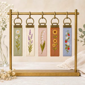 Puede incluir: Cinco llaveros de tela rectangulares con diseños botánicos bordados colgados de un soporte de latón. Los diseños presentan una margarita, lavanda, lirio de los valles, girasol y una planta de fresa sobre tela color salvia, crema, rosa, naranja y azul.