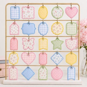 May include: A gold-toned metal display rack holds twenty-five assorted fabric tags in various shapes, including hearts, stars, butterflies, and rectangles. The tags feature pastel colors, gingham patterns, polka dots, and floral prints with stitched edges.