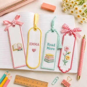 May include: Four white fabric bookmarks with embroidered designs and ribbon accents. One features an open book, one says EMMA with a heart, one says Read More with a stack of books, and one shows flowers. Each has a distinct border and top ribbon.